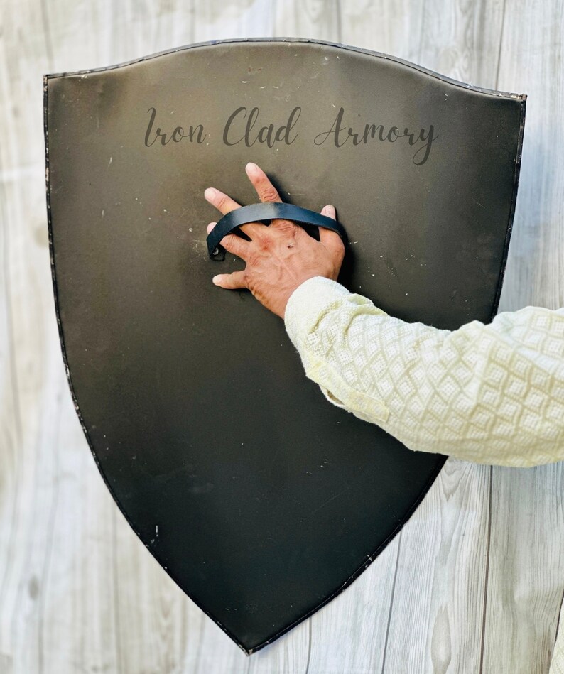 May include: A black, shield-shaped object with a leather handle. The shield is made of metal and has the words "Iron Clad Armory" printed on it.