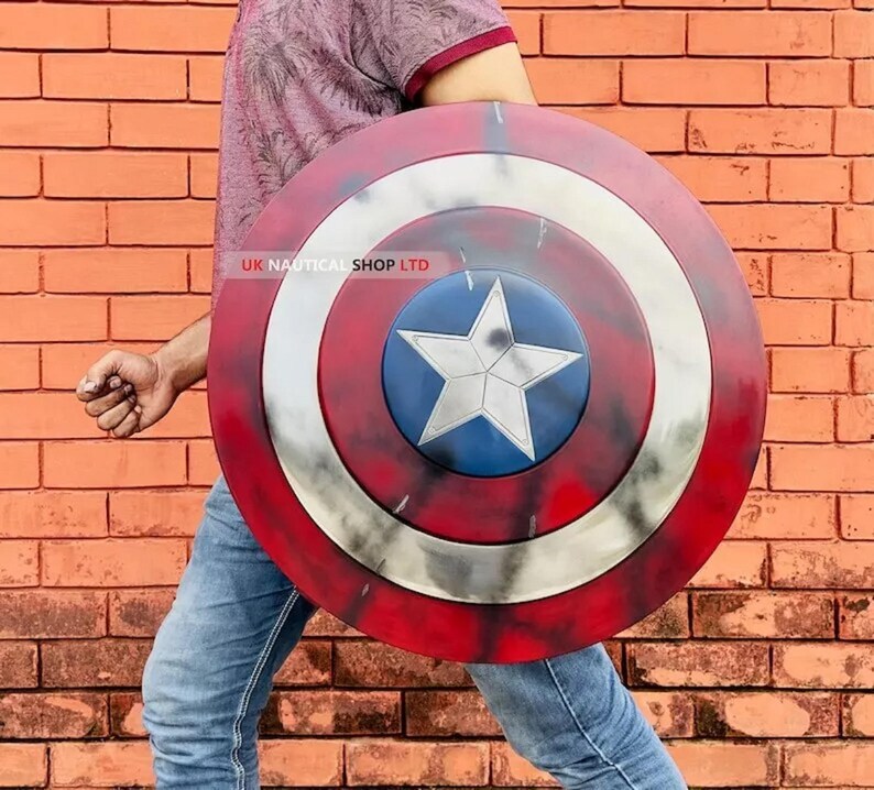 May include: A red, white, and blue Captain America shield with a five-pointed star in the center. The shield is made of metal and has a distressed look. UK Nautical Shop LTD is printed on the shield.