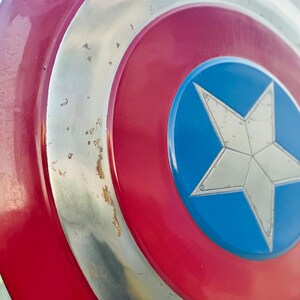 K&ouml;nnte beinhalten: Ein rot-wei&szlig;-blauer Captain America Schild mit einem silbernen Stern in der Mitte. Der Schild hat einen abgenutzten, verwitterten Look mit einigen Rostflecken auf dem Metall.