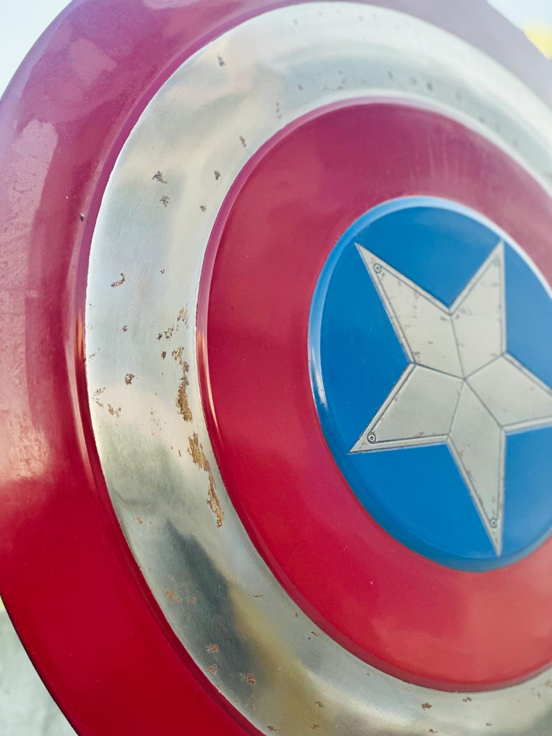 K&ouml;nnte beinhalten: Ein rot-wei&szlig;-blauer Captain America Schild mit einem silbernen Stern in der Mitte. Der Schild hat einen abgenutzten, verwitterten Look mit einigen Rostflecken auf dem Metall.
