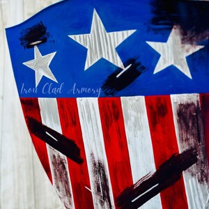 May include: A blue and red shield with white stars and stripes. The shield has a distressed look with black paint splatters. The text "Iron Clad Armory" is visible at the bottom of the shield.