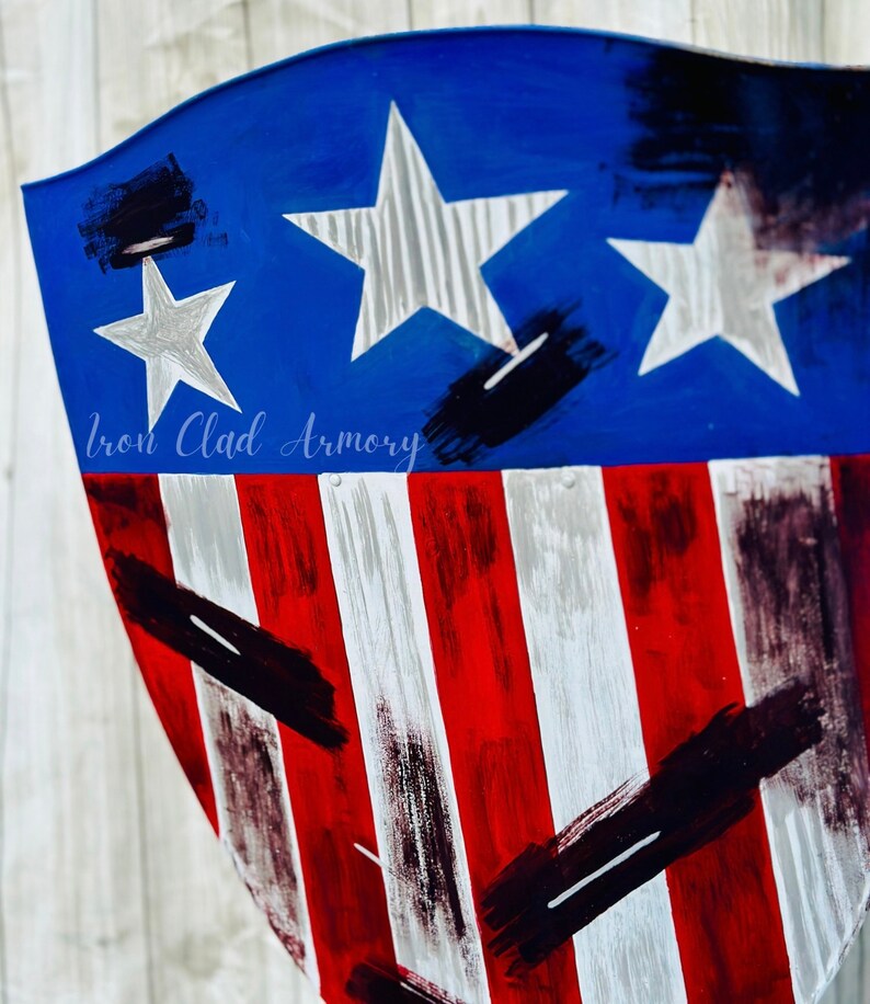 May include: A blue and red shield with white stars and stripes. The shield has a distressed look with black paint splatters. The text "Iron Clad Armory" is visible at the bottom of the shield.