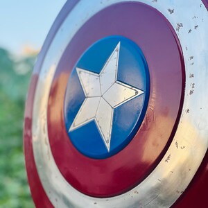 K&ouml;nnte beinhalten: Ein rot-wei&szlig;-blauer Captain America Schild mit einem gro&szlig;en, f&uuml;nfzackigen Stern in der Mitte. Der Schild hat eine metallische Oberfl&auml;che und zeigt Gebrauchsspuren.