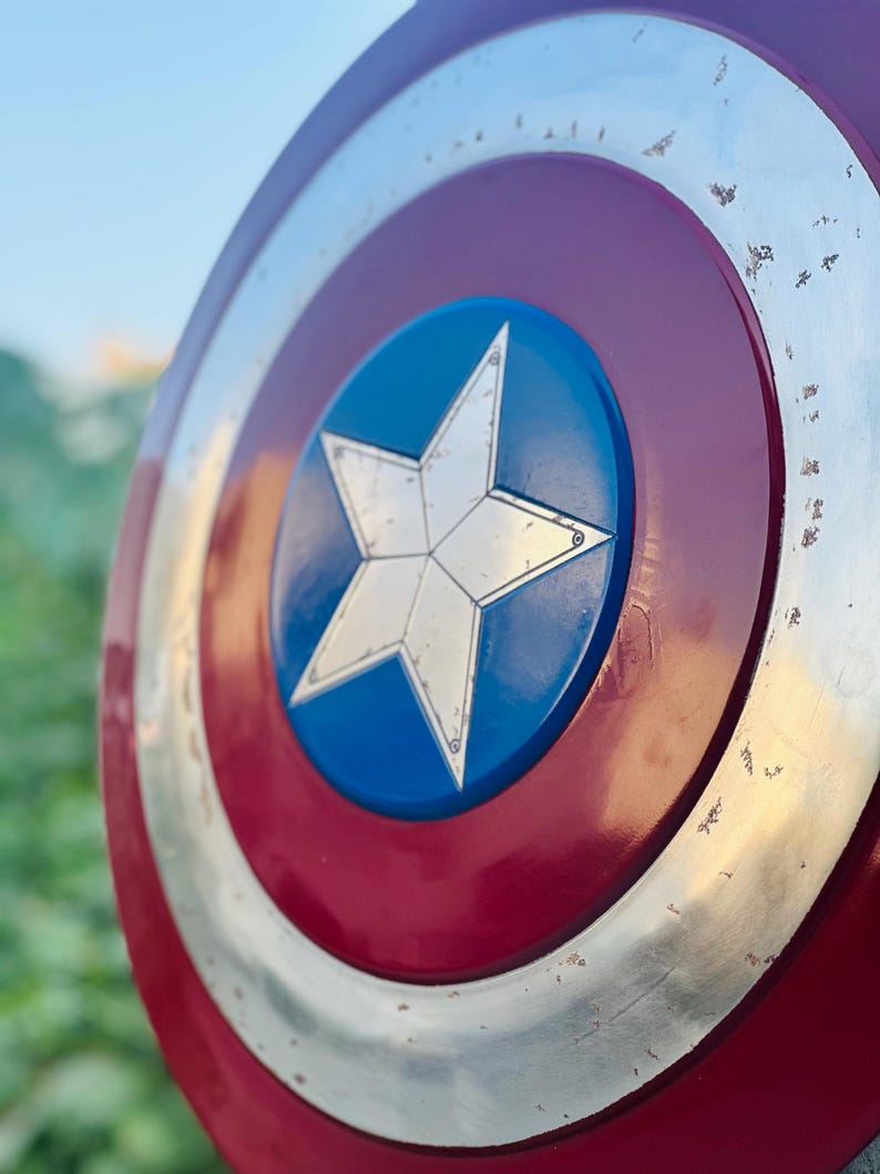 K&ouml;nnte beinhalten: Ein rot-wei&szlig;-blauer Captain America Schild mit einem gro&szlig;en, f&uuml;nfzackigen Stern in der Mitte. Der Schild hat eine metallische Oberfl&auml;che und zeigt Gebrauchsspuren.