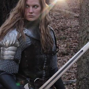 May include: A woman in a forest setting, wearing chainmail armor and a leather tunic, holds a sword in her right hand. She has long, brown hair and is looking off to the side.
