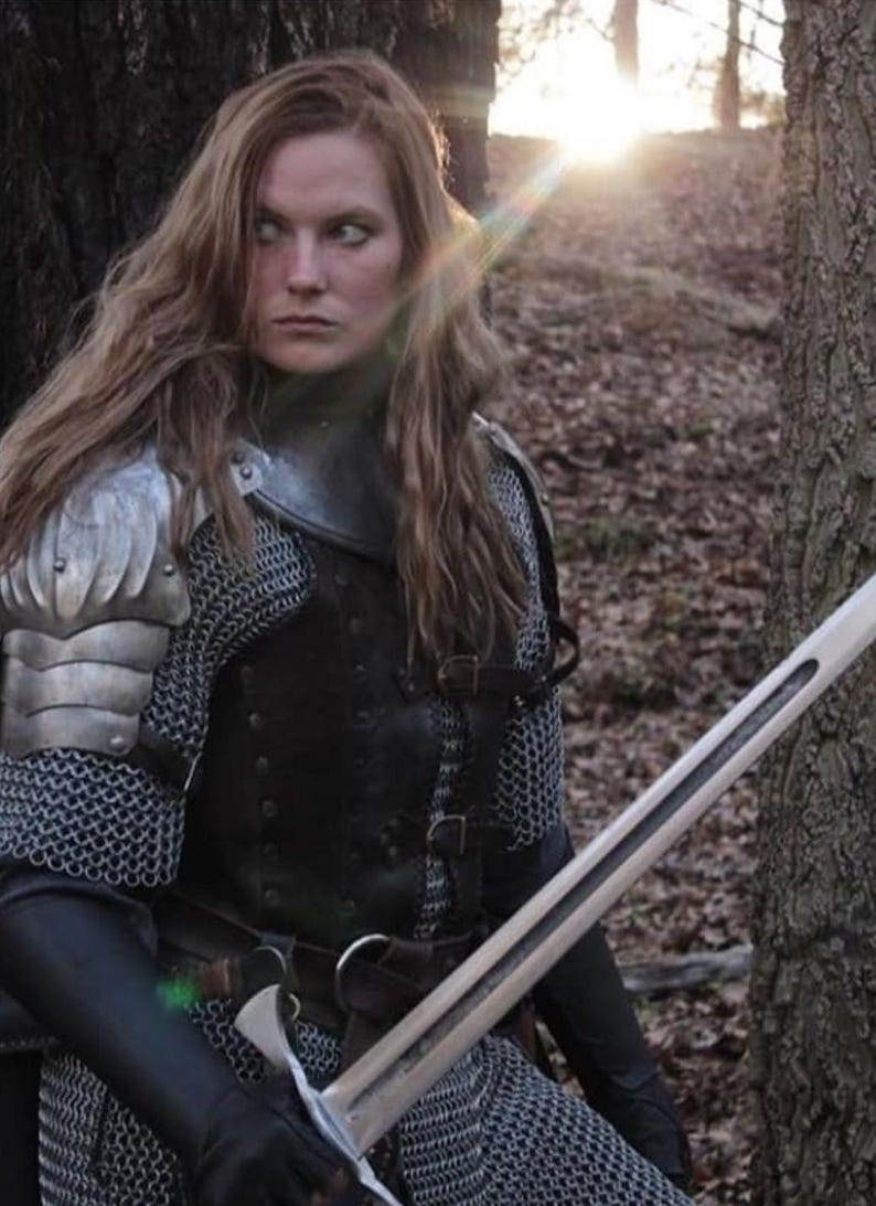 May include: A woman in a forest setting, wearing chainmail armor and a leather tunic, holds a sword in her right hand. She has long, brown hair and is looking off to the side.