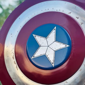 K&ouml;nnte beinhalten: Ein rot-wei&szlig;-blauer Captain-America-Schild mit einem gro&szlig;en, wei&szlig;en f&uuml;nfzackigen Stern in der Mitte. Der Schild hat einen silbernen Au&szlig;enrand und einen abgenutzten, verwitterten Look.
