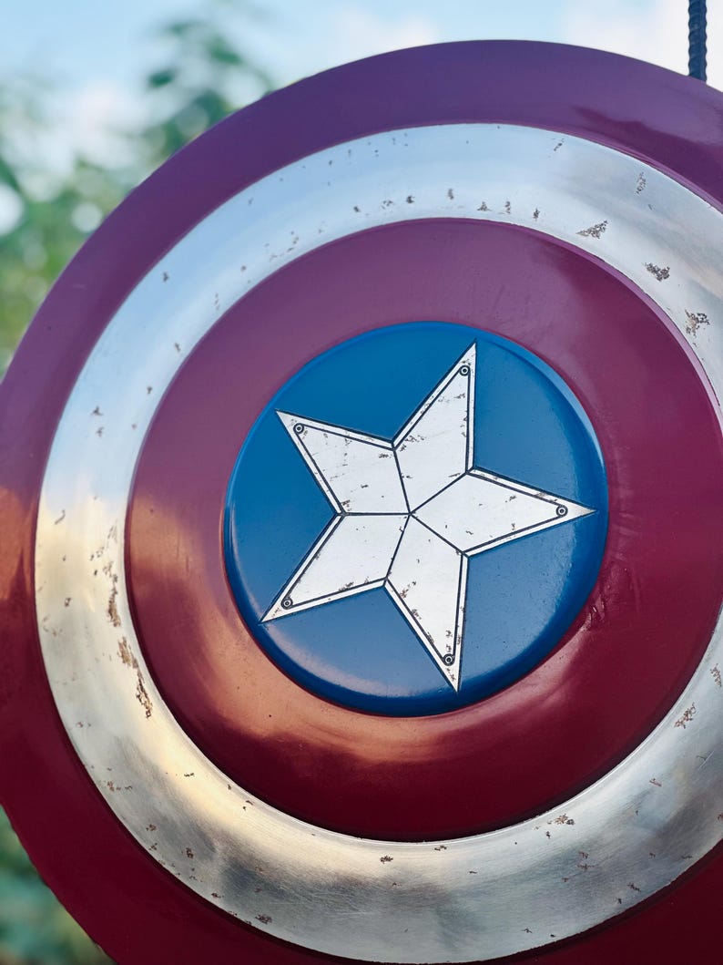 K&ouml;nnte beinhalten: Ein rot-wei&szlig;-blauer Captain-America-Schild mit einem gro&szlig;en, wei&szlig;en f&uuml;nfzackigen Stern in der Mitte. Der Schild hat einen silbernen Au&szlig;enrand und einen abgenutzten, verwitterten Look.