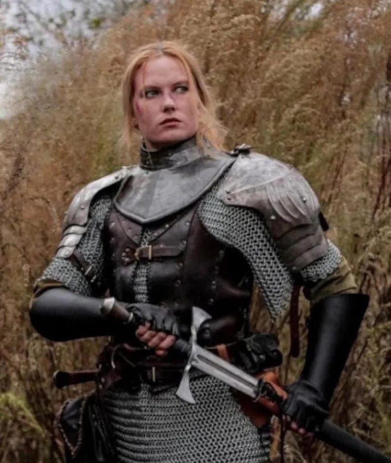 May include: A woman in medieval armor, wearing a chainmail shirt, leather armor, and a helmet. She is holding a sword in her right hand and looking to the left.