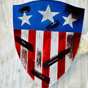 May include: A red, white, and blue shield with a distressed paint finish. The shield has a blue background with three white stars and red and white stripes. The shield is painted with black brushstrokes and the text "Iron Clad Armory" is written in black.