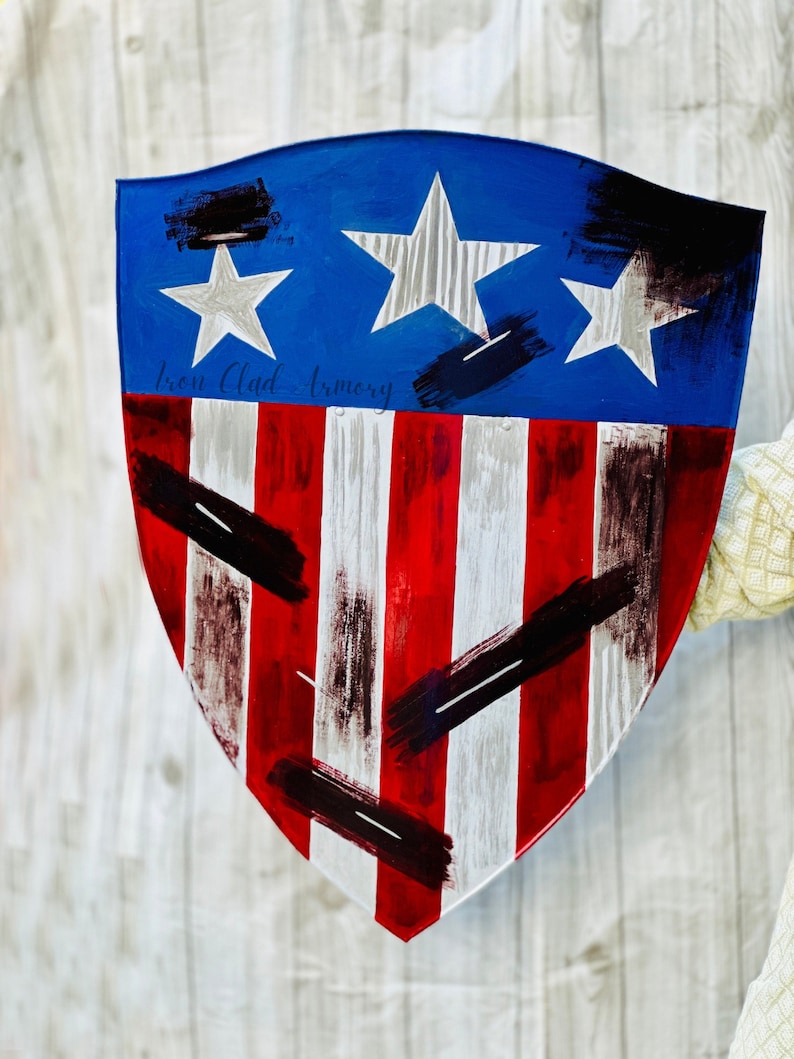May include: A red, white, and blue shield with a distressed paint finish. The shield has a blue background with three white stars and red and white stripes. The shield is painted with black brushstrokes and the text "Iron Clad Armory" is written in black.