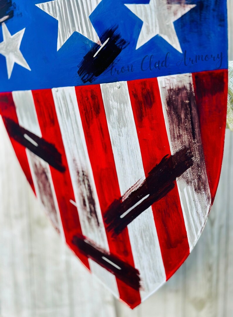 May include: A red, white, and blue shield with black paint splatters. The shield is shaped like a heart and has the text "Iron Clad Armory" written on it.