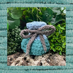 Könnte beinhalten: Ein handgefertigter Häkelbeutel in Blau- und Türkistönen, gebunden mit einer grauen Schleife. Der Beutel ist aus weichem, strukturiertem Garn gefertigt und liegt auf einem Holzstück, umgeben von Grün. Ein bezaubernder, kleiner und dekorativer Artikel.