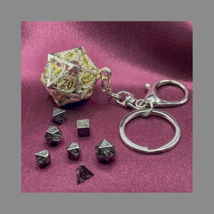 Könnte beinhalten: Ein silberfarbener, verzierter D20-Würfel-Schlüsselanhänger mit einem passenden Satz polyedrischer Würfel. Der große Würfel ist mit goldenen Akzenten und der Zahl 20 versehen. Die Würfel liegen auf einem bordeauxfarbenen Stoff.