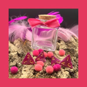 Op de afbeelding: Een set roze en gouden driehoekige dobbelstenen met de nummers 1, 2, 3 en 4. De dobbelstenen liggen op een houten oppervlak met roze pompoenen en een klein glazen potje met een kurk en een roze lint.