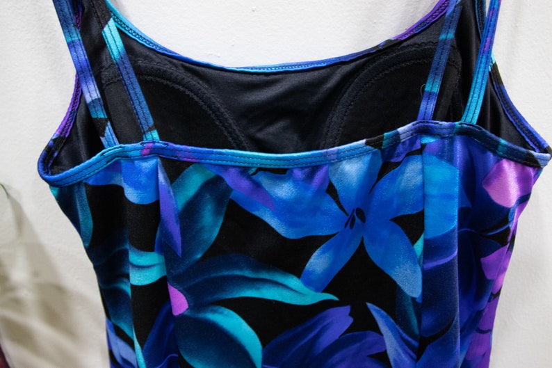 Puede incluir: Un ba&ntilde;ador negro con estampado floral azul y morado. El ba&ntilde;ador tiene tirantes finos y un escote redondo.