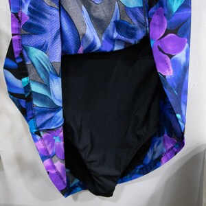 Puede incluir: Un traje de ba&ntilde;o negro con estampado floral en tonos azules, morados y turquesa. El traje de ba&ntilde;o tiene un escote redondo y un estilo de espalda de nadador.
