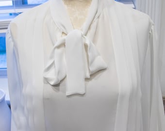 Puede incluir: Una blusa blanca de manga larga con un lazo en el cuello.
