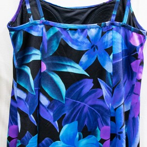 Puede incluir: Un traje de ba&ntilde;o negro con estampado floral azul y morado. El traje de ba&ntilde;o tiene tirantes ajustables y un escote redondo.