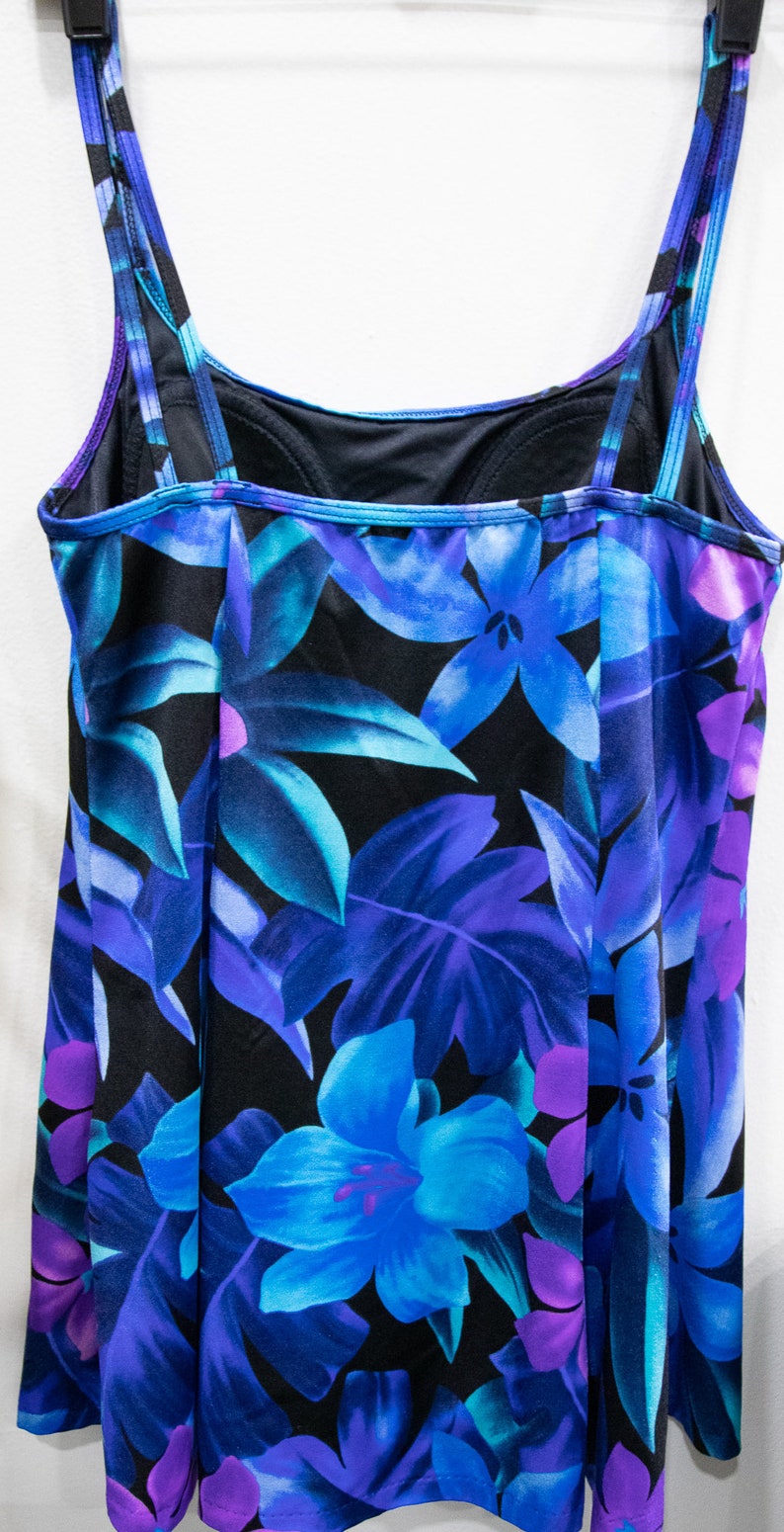Puede incluir: Un traje de ba&ntilde;o negro con estampado floral azul y morado. El traje de ba&ntilde;o tiene tirantes ajustables y un escote redondo.