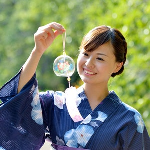 Pode incluir: Uma mulher vestindo um quimono azul floral segura um sino de vento de vidro transparente com um design floral. O sino de vento é suspenso por uma corda. O fundo é verde desfocado, sugerindo um ambiente ao ar livre.
