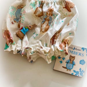 May include: Cream-coloured baby bloomers with a ruffle trim, featuring a Peter Rabbit print with bunnies and floral accents. A small Peter Rabbit figurine and a "Peter Rabbit Numbers" book are also in the image.