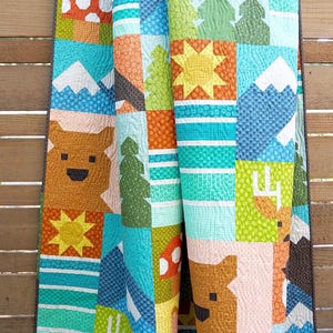 May include: A patchwork quilt featuring a repeating nature-themed pattern with blocks of brown bears, yellow suns, green pine trees, white snow-capped mountains, and blue striped sections. The fabric displays various textures and geometric shapes.