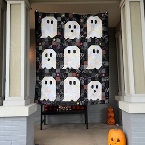 May include: A rectangular quilt featuring a grid of nine white ghost shapes appliqued onto a black background. The ghosts have dark oval eyes and mouths, and the black fabric blocks are patterned with small white and orange geometric designs.
