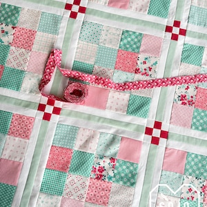 May include: A patchwork quilt in shades of pink, mint green, and white, with a decorative pink ribbon. The quilt features a grid pattern of squares with floral and geometric designs. Red squares accent the intersections.