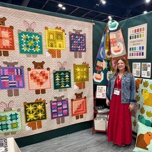 May include: A large quilt featuring repeating animal figures with geometric patterns in various colors. The quilt is displayed on a wall, with a woman in a red dress standing nearby. Additional quilts and color charts are also visible.