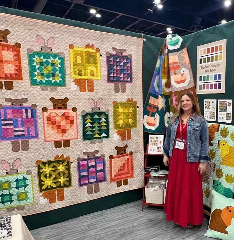 May include: A large quilt featuring repeating animal figures with geometric patterns in various colors. The quilt is displayed on a wall, with a woman in a red dress standing nearby. Additional quilts and color charts are also visible.