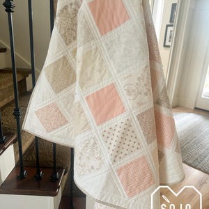 May include: A handmade quilt with a patchwork design. The quilt features squares of various patterns and colors, including peach, beige, and white. The quilt is draped over a staircase, showcasing its size and design.