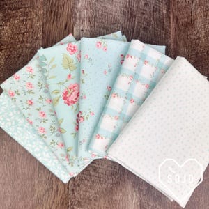 May include: A collection of folded fabric squares in various floral and patterned designs. The fabrics feature pastel colors like light blue and white, with pink and red floral accents. The fabrics are arranged on a wooden surface.