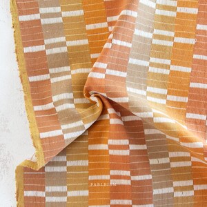 May include: Close-up of a folded textile with a geometric pattern. The fabric features alternating vertical stripes of orange, white, and tan rectangles. The edge of the fabric has a yellow border. The word "FABLEISM" is visible.