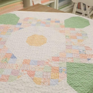 May include: A white quilt with a large flower design in the center. The flower is made up of patchwork squares in various shades of pink, blue, green, and yellow. The center of the flower is a yellow circle with a checkered pattern.