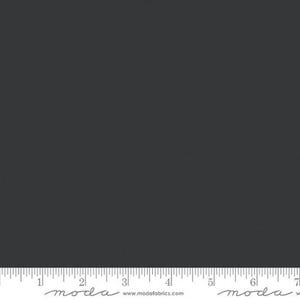 May include: A solid charcoal gray fabric swatch featuring a smooth, uniform texture. At the bottom edge, a white ruler is visible, showing measurements in inches from 0 to 7, with the text "moda" and "www.modafabrics.com" printed clearly.