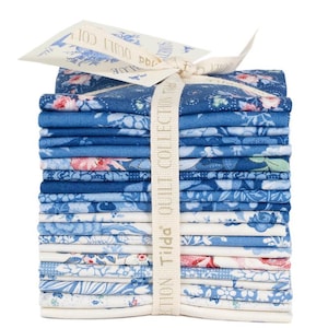 May include: A stack of blue fabric squares with floral patterns, tied with a cream ribbon and tag. The fabric features various shades of blue and white with pink accents. The tag reads "Tilda Quilt Collection".