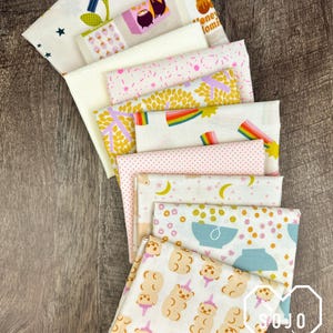 May include: A collection of ten folded fabric squares featuring various patterns including a blue bird, honey pots, pink strawberries, yellow leaves, rainbows, pink polka dots, yellow moons, cereal bowls, and light brown gummy bears with party hats.