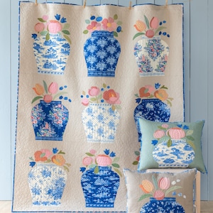 May include: A beige quilt and two throw pillows feature a repeating pattern of blue and white vases filled with colorful flowers. The quilt is hanging on a light blue wall. The pillows are in shades of blue and beige.