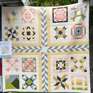 May include: A large, handmade quilt with a patchwork design. The quilt features various geometric patterns in pastel colors, including pink, yellow, and blue, set against a white background. The quilt is displayed outdoors.