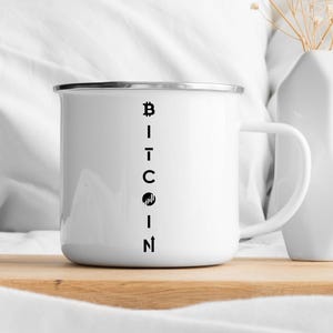 May include: White enamel mug with a black Bitcoin design. The mug features the word "Bitcoin" vertically aligned on the front. The mug has a silver rim and a white handle. The mug is sitting on a wooden surface.