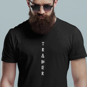 Pode incluir: Uma camiseta preta com um gráfico vertical branco que lê "TRAINER" com letras estilizadas.
