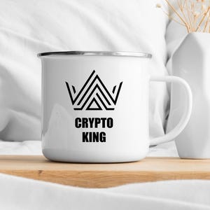 May include: White enamel mug with a black geometric crown design and the text "CRYPTO KING". The mug has a silver rim and a white handle. The mug is sitting on a wooden surface.