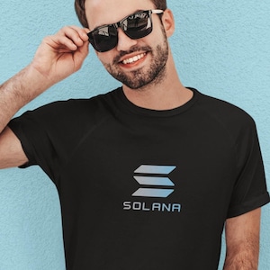 May include: A man wearing a black t-shirt with a blue and white logo that says "SOLANA". He is wearing sunglasses and has a beard.