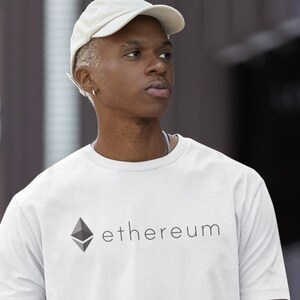 May include: White t-shirt with the word "ethereum" printed in black. The shirt features a geometric logo above the text. The person in the image is wearing a white cap and black trousers. The shirt is a crew neck style.