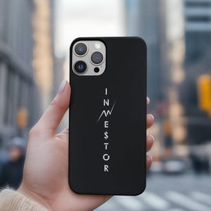 May include: Black phone case with the word "INVESTOR" in white, vertically aligned. The design includes a stylized upward arrow and a dollar sign. The phone case is held in a hand, with a blurred city background.
