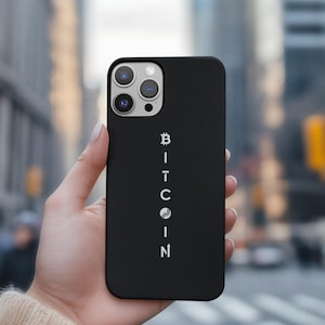 May include: Black phone case with the word "BITCOIN" printed vertically in white. The case has a sleek design and is held in a hand. The background is blurred, showing a city scene.