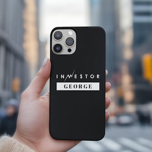 May include: Black phone case with the text "INVESTOR GEORGE" in white. The phone case is held in a hand against a blurred city background. The phone case has a camera module with three lenses.