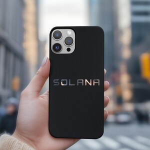 May include: Black phone case with the word "SOLANA" in silver lettering. The phone case has a camera module with three lenses. The background is a blurred city scene.