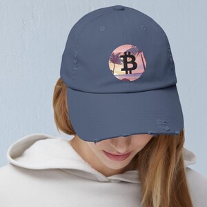 May include: A blue baseball cap with a distressed brim. The cap features a circular design with a black Bitcoin symbol set against a sunset scene with palm trees.